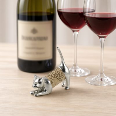 Lucina K Cat Cork Pet, a solid pewter stretching cat with its tail held high holding a wine cork, beside a wine bottle and two glasses of red wine