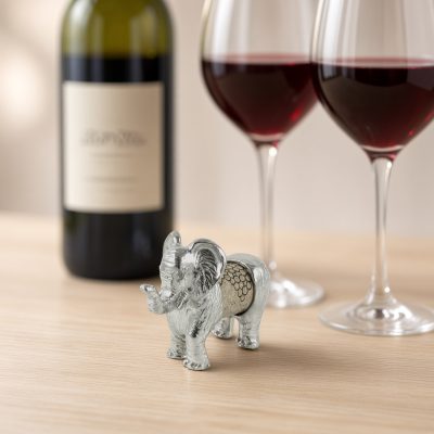 Lucina K Elephant Cork Pet, a solid pewter elephant with a raised trunk holding a wine cork, beside a wine bottle and two glasses of red wine