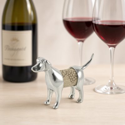 Lucina K Lab Cork Pet, a solid pewter Labrador Retriever with a long tail holding a wine cork, beside a wine bottle and two glasses of red wine