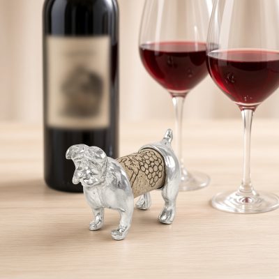 Lucina K Bulldog Cork Pet, a solid pewter stocky Bulldog with a wrinkled face holding a wine cork, beside a wine bottle and two glasses of red wine