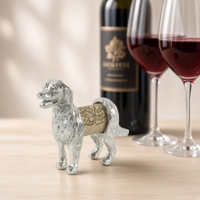 Lucina K Retriever Cork Pet, a solid pewter Golden Retriever with a feathered coat holding a wine cork, beside a wine bottle and two glasses of red wine