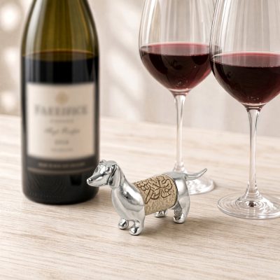 Lucina K Dachshund Cork Pet, a solid pewter long-bodied Dachshund holding a wine cork, beside a wine bottle and two glasses of red wine