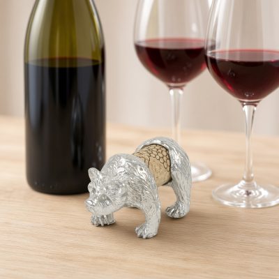 Lucina K Bear Cork Pet, a solid pewter textured walking bear holding a wine cork, beside a wine bottle and two glasses of red wine