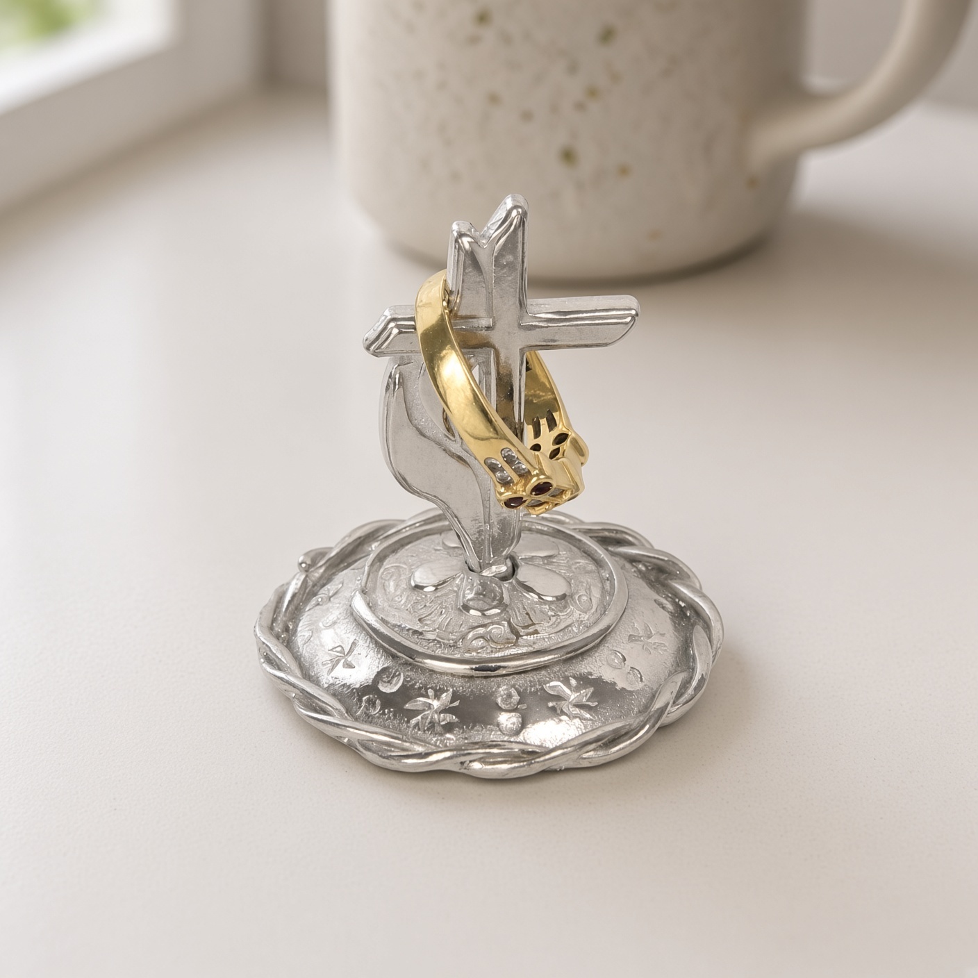 Pewter cross and flame ring stand with a gold ring hanging on the cross arm on a windowsill beside a speckled mug, ring not included