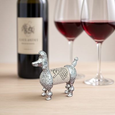 Lucina K Poodle Cork Pet, a solid pewter Poodle with a pom-pom clipped coat holding a wine cork, beside a wine bottle and two glasses of red wine