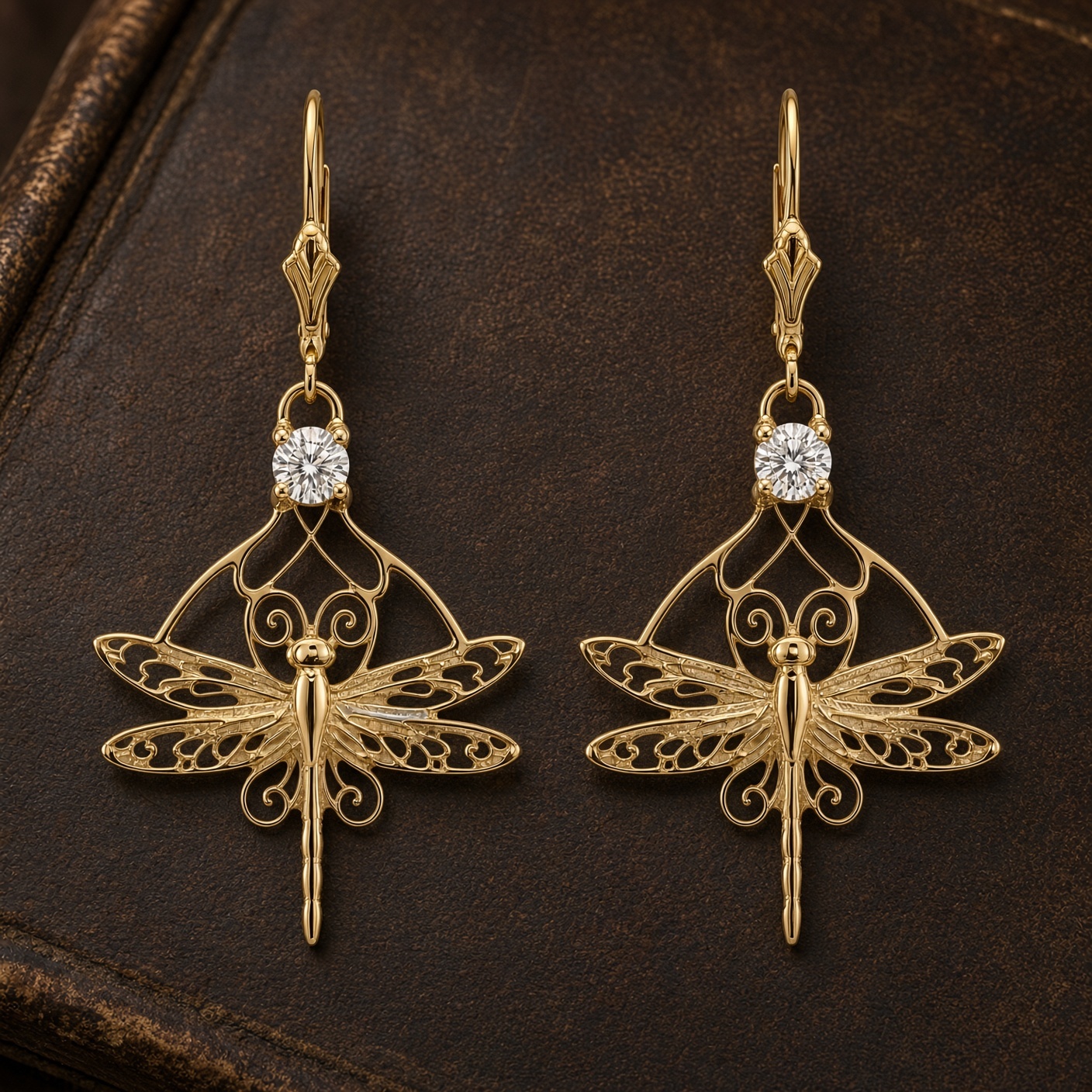 Pair of 14K gold filigree dragonfly drop earrings with round diamonds on a dark brown leather surface